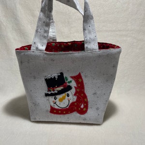 Op de afbeelding: Een witte canvas draagtas met een sneeuwmanontwerp. De tas heeft een rood-wit sneeuwvlokkenpatroon en een rode voering. De sneeuwman draagt ​​een zwarte hoge hoed en een rode sjaal met gouden sterren.