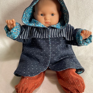 Puede incluir: Una muñeca con una chaqueta con capucha azul marino con detalles de rayas blancas y un forro con estampado floral. La muñeca también lleva pantalones color óxido con motas doradas y zapatos dorados. La chaqueta tiene un diseño acolchado con pequeños patrones blancos en forma de estrella.