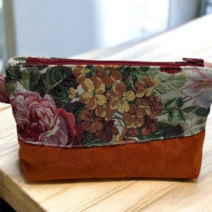 Puede incluir: Un neceser rectangular con una parte superior de tapiz floral y una sección inferior de ante color óxido. La bolsa tiene una cremallera burdeos y un pequeño lazo en un lado. El estampado floral incluye tonos rojos, rosas, amarillos y verdes.