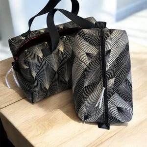 Peut inclure: Ensemble de deux sacs de voyage. Les sacs présentent un motif géométrique noir et or. Le plus grand sac a une poignée noire et un intérieur rouge. Le plus petit sac a une fermeture éclair. Les deux sacs sont sur une surface en bois clair.