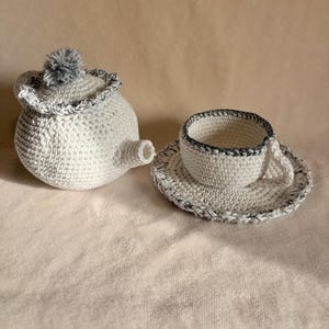 Könnte beinhalten: Ein gehäkeltes Teeservice in Weiß mit grauen Akzenten. Das Set besteht aus einer Teekanne mit Bommel auf dem Deckel, einer Teetasse und einer Untertasse. Die Ränder von Teekanne, Tasse und Untertasse sind mit grauem Garn verziert.