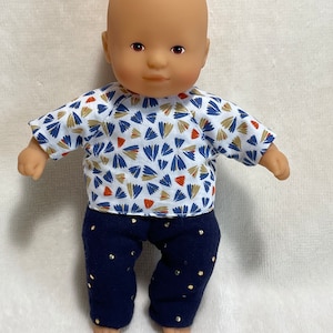 Peut inclure: Un poupon chauve portant une chemise à motifs bleus et blancs avec des accents orange et un pantalon bleu marine à pois dorés.