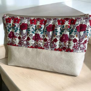 Puede incluir: Una bolsa de cremallera beige con estampado floral. La mitad superior de la bolsa es de tela crema con diseños florales rojos, rosas y verdes. La mitad inferior es de tela beige lisa.