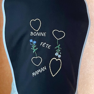 以下が含まれることがあります： 白い縁取りと刺繍されたハート、花、そして「Bonne Fête Maman」の文字が書かれた黒いよだれかけ。