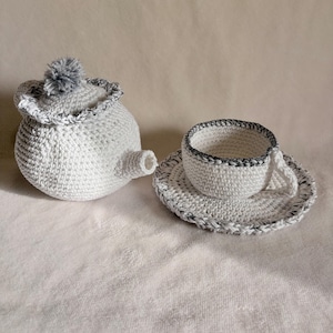 May include: A white crocheted tea set, including a teapot with a gray pom-pom on the lid, a teacup, and a saucer. The edges of the set are trimmed with a black and white pattern. The set is handmade.
