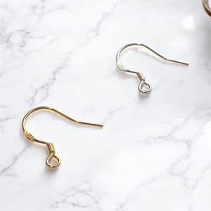 May include: Two earring hooks, one gold-colored and one silver-colored, are displayed against a white marble background. The hooks are curved with a loop at the bottom for attaching earrings.