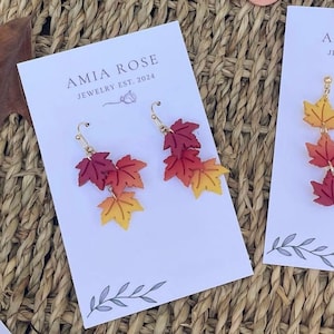 May include: A pair of leaf-shaped earrings with a gradient of colors from red to yellow. The earrings are on a white card with the text "AMIA ROSE JEWELRY EST. 2024". The earrings are hanging on gold-colored hooks.