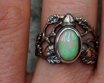 May include: A silver ring with an oval-shaped opal gemstone set in a delicate, leafy design. The ring features a branch-like setting with acorns on either side of the stone.