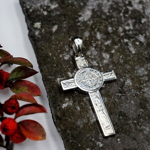 May include: A silver pendant in the shape of a cross with intricate details. The center of the cross features a round medallion with a detailed design.