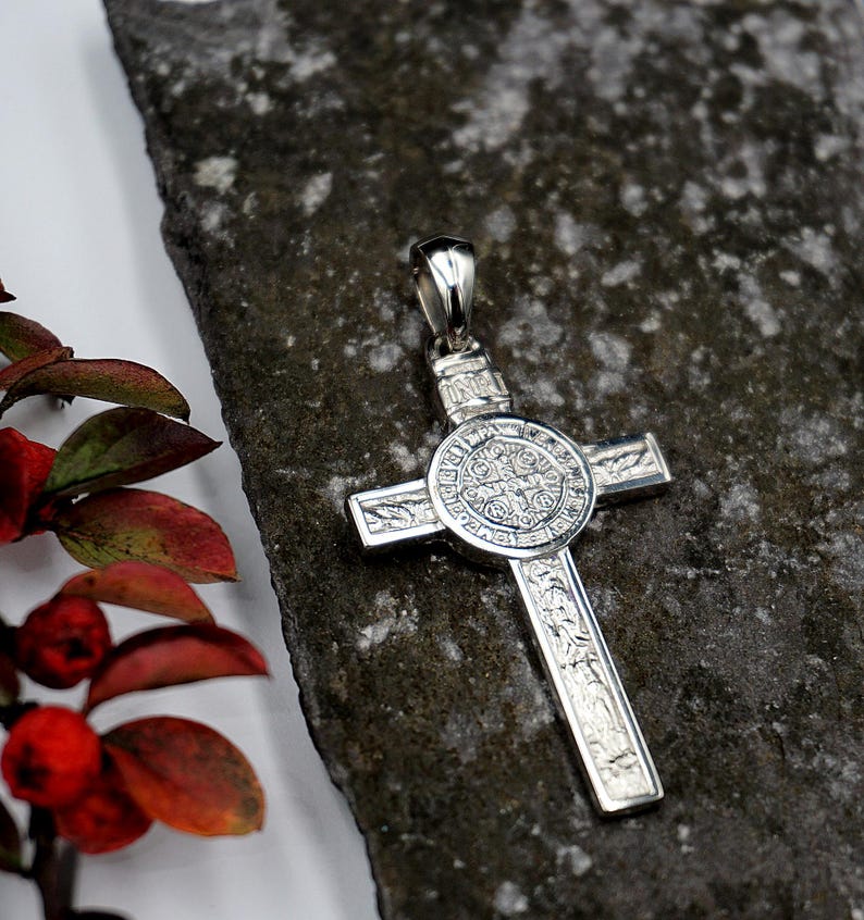 May include: A silver pendant in the shape of a cross with intricate details. The center of the cross features a round medallion with a detailed design.