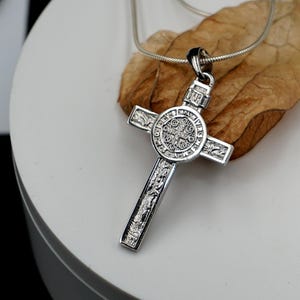 May include: A silver chain necklace with a detailed silver cross pendant. The cross features a circular design with intricate engravings.