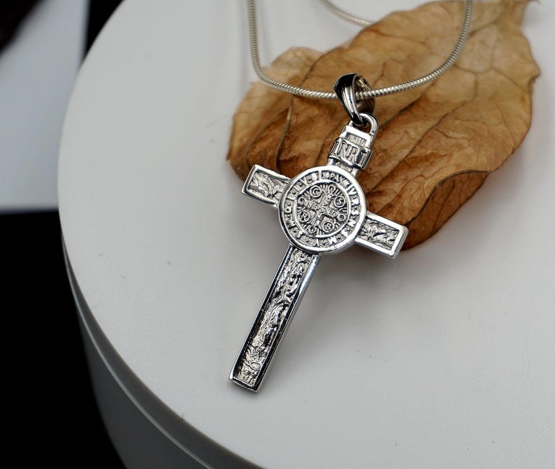 May include: A silver chain necklace with a detailed silver cross pendant. The cross features a circular design with intricate engravings.