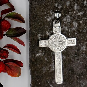 May include: A silver St. Benedict's medal pendant in the shape of a cross. The medal features a detailed design with the words "INRI" and "ORA PRO NOBIS" inscribed on it.
