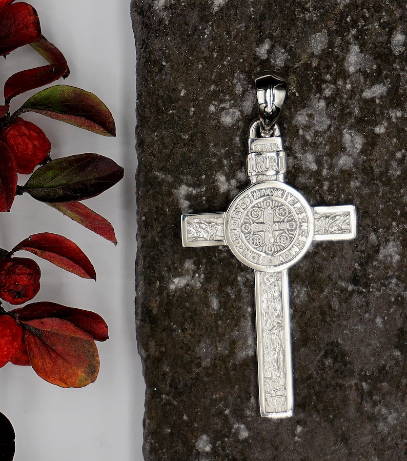 May include: A silver St. Benedict's medal pendant in the shape of a cross. The medal features a detailed design with the words "INRI" and "ORA PRO NOBIS" inscribed on it.