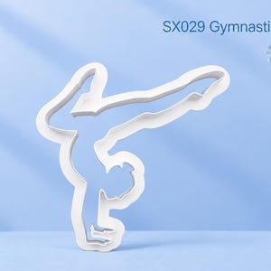 Op de afbeelding: Een witte koekjesvorm in de vorm van een turner die een handstand maakt, met de tekst SX029 Gymnastics Handstand. De omtrek toont een figuur met armen naar beneden gestrekt en benen in een spagaat, gemaakt van stevig wit materiaal.