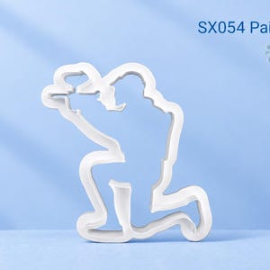 Könnte beinhalten: Ein weißer Kunststoff-Ausstecher in Form eines knienden Paintballspielers, der einen Markierer hält. Der Text SX054 Paintball Player ist in der oberen rechten Ecke aufgedruckt und ein gelbes Logo ist in der oberen linken Ecke zu sehen.