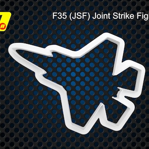 May include: White plastic cookie cutter shaped like an F-35 Joint Strike Fighter aircraft. The cutter is on a black and blue background.