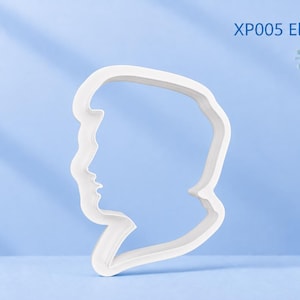 Puede incluir: Un cortador de galletas blanco con forma de perfil lateral de una persona con peinado pompadour, etiquetado como XP005 Elvis Greek God. El objeto es un contorno blanco sólido de una cabeza y cuello, diseñado para cortar masa en una silueta.