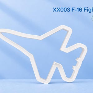 Può includere: Un tagliabiscotti in plastica bianca a forma di aereo F-16 Fighting Falcon. Il testo XX003 F-16 Fighting Falcon è stampato in blu nell'angolo in alto a destra. L'oggetto presenta un contorno netto e angolare del caccia militare.