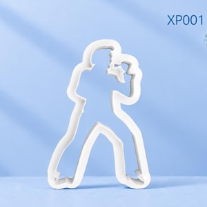 Puede incluir: Un cortador de galletas blanco con la silueta de una persona cantando en un micrófono, con el texto XP001 Elvis Presley impreso en la esquina superior derecha. El objeto tiene un centro hueco y un borde elevado para cortar la masa.
