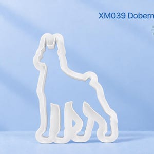 May include: A white Dobermann Pinscher cookie cutter with a distinct dog silhouette outline. The product is labelled with the text XM039 Dobermann Pinscher in the upper right corner. The cutter features a clean, minimalist design for shaping dough.