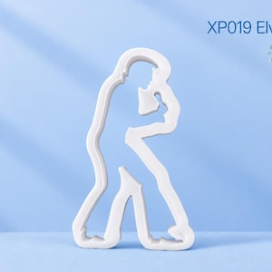 Puede incluir: Un cortador de galletas blanco con forma de cantante sosteniendo un micrófono, con el texto XP019 Elvis In Concert. El contorno muestra a una persona en una postura dinámica con las piernas abiertas, capturando una pose de actuación en blanco.