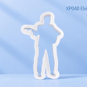 Puede incluir: Un cortador de galletas blanco con la forma de una figura de pie tocando la guitarra, con el texto XP048 Elvis Guitar Player impreso en la esquina superior derecha. El objeto presenta un contorno de plástico moldeado liso de un músico con instrumento.