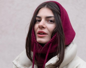 May include: A deep magenta knitted balaclava hood with a ribbed texture and long matching drawstrings, worn over a cream-colored puffer jacket with a high collar and snap button closures.