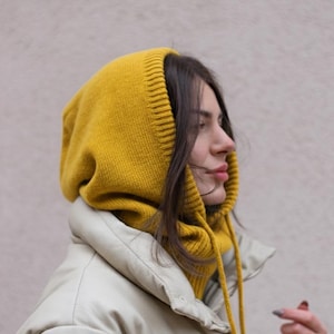 Könnte beinhalten: Eine senfgelbe Strick-Balaclava mit Kordelzug, getragen über einer cremefarbenen Pufferjacke. Die Balaclava bedeckt Kopf und Hals, das Gesicht ist sichtbar. Die Jacke hat ein glattes, mattes Finish.