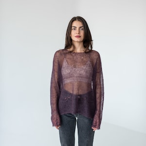 Può includere: Una donna indossa un maglione viola trasparente sopra un reggiseno color carne e jeans scuri.