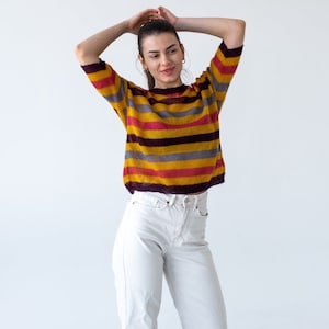 May include: A woman wearing a yellow, pink, and purple striped sweater with white jeans.
