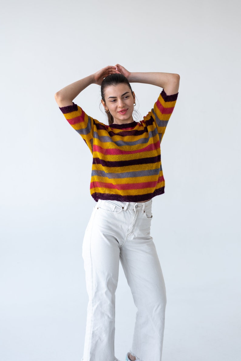 May include: A woman wearing a yellow, pink, and purple striped sweater with white jeans.
