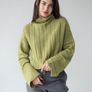 May include: A woman wearing a light green ribbed knit sweater with a turtleneck and flared sleeves. The sweater is paired with gray trousers. The background is a plain, light gray.