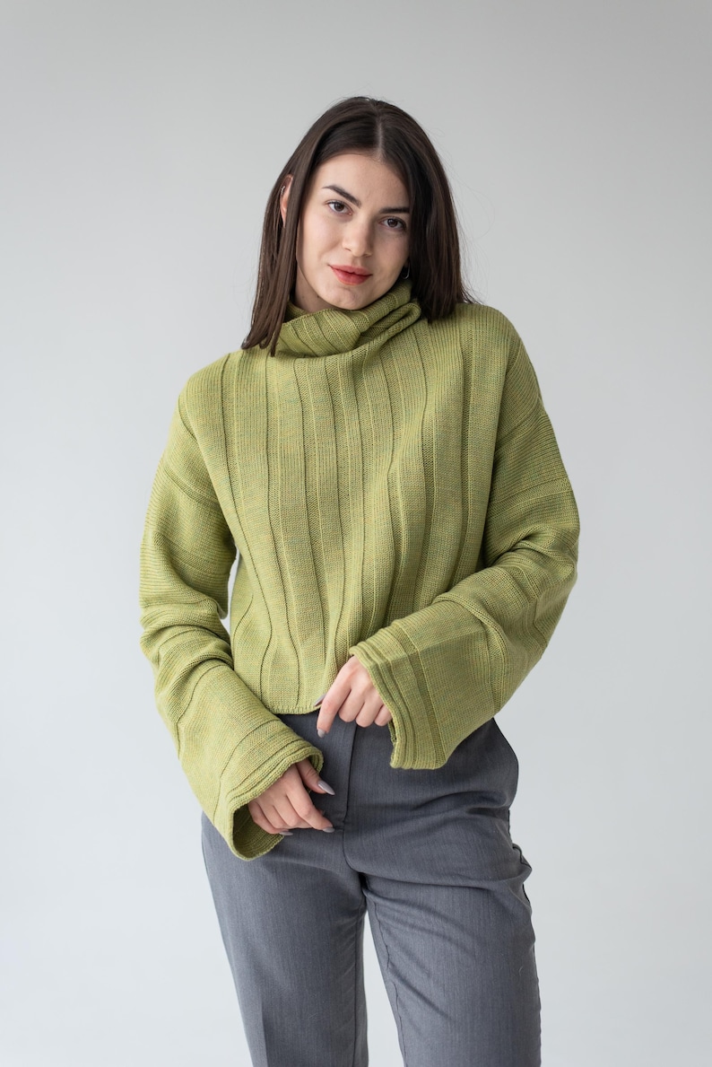 May include: A woman wearing a light green ribbed knit sweater with a turtleneck and flared sleeves. The sweater is paired with gray trousers. The background is a plain, light gray.