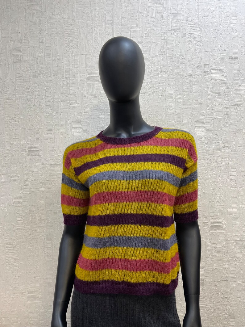 May include: A striped short-sleeved sweater in shades of yellow, purple, pink, and gray. The sweater is made of a soft, fuzzy material.