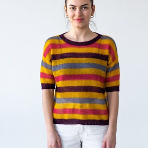 May include: A woman wearing a striped sweater with yellow, red, purple, and gray stripes. She is wearing white pants and has her hair pulled back in a bun.