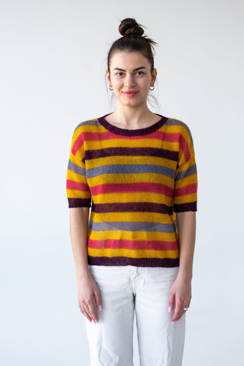 May include: A woman wearing a striped sweater with yellow, red, purple, and gray stripes. She is wearing white pants and has her hair pulled back in a bun.