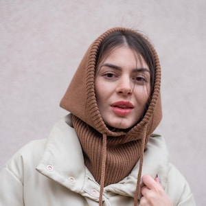 Puede incluir: Una mujer con una bufanda de capucha de punto marr&oacute;n sobre una chaqueta acolchada de color crema.