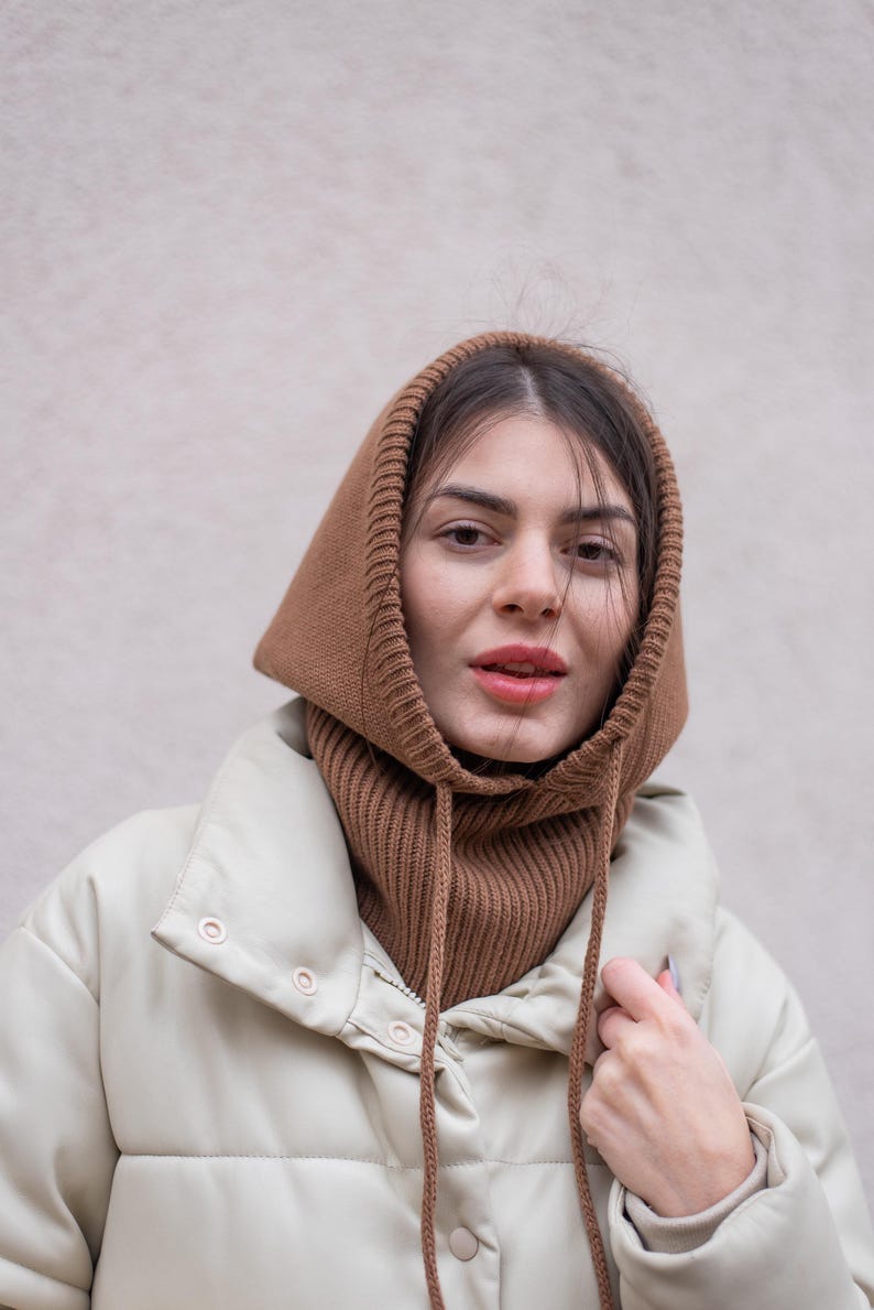 Puede incluir: Una mujer con una bufanda de capucha de punto marr&oacute;n sobre una chaqueta acolchada de color crema.