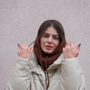 Puede incluir: Una mujer con un cuello de punto marr&oacute;n y una chaqueta acolchada de color crema. Est&aacute; ajustando el cuello con las manos.