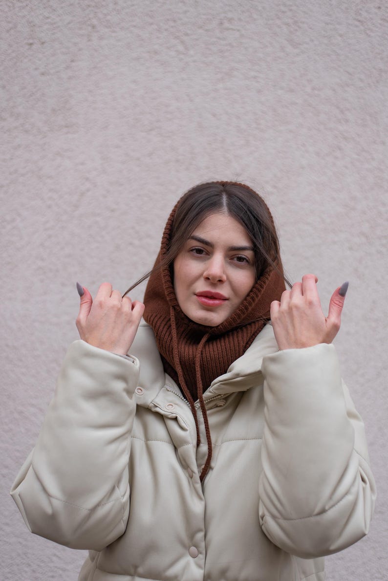 Puede incluir: Una mujer con un cuello de punto marr&oacute;n y una chaqueta acolchada de color crema. Est&aacute; ajustando el cuello con las manos.
