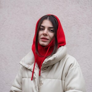 Puede incluir: Una mujer con una chaqueta acolchada blanca y una bufanda de punto roja con capucha. La bufanda tiene un cord&oacute;n.