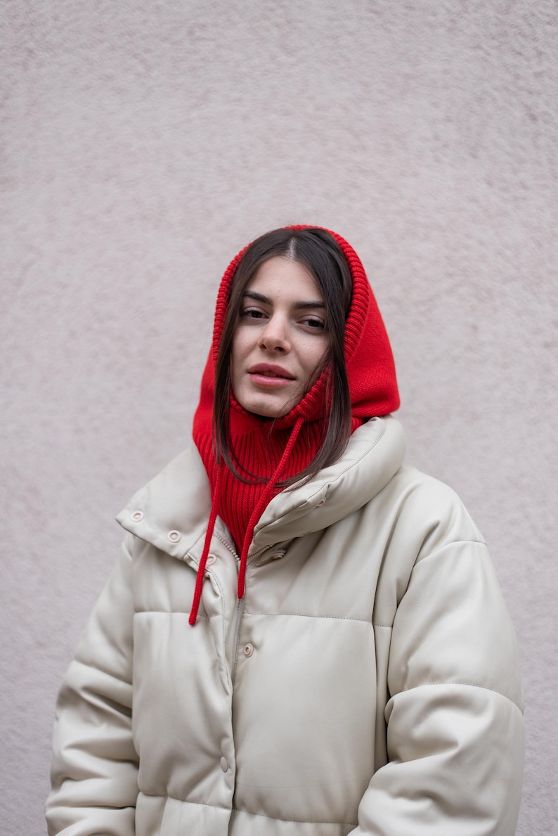 Puede incluir: Una mujer con una chaqueta acolchada blanca y una bufanda de punto roja con capucha. La bufanda tiene un cord&oacute;n.