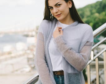 May include: A woman wearing a light gray cardigan over a white t-shirt and blue denim shorts. She is standing on a staircase with a metal railing.