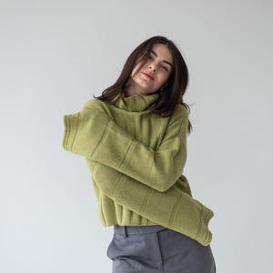 May include: A woman wearing a light green ribbed knit turtleneck sweater and gray trousers. The sweater has long sleeves and a high neckline. The pants are a solid gray color.
