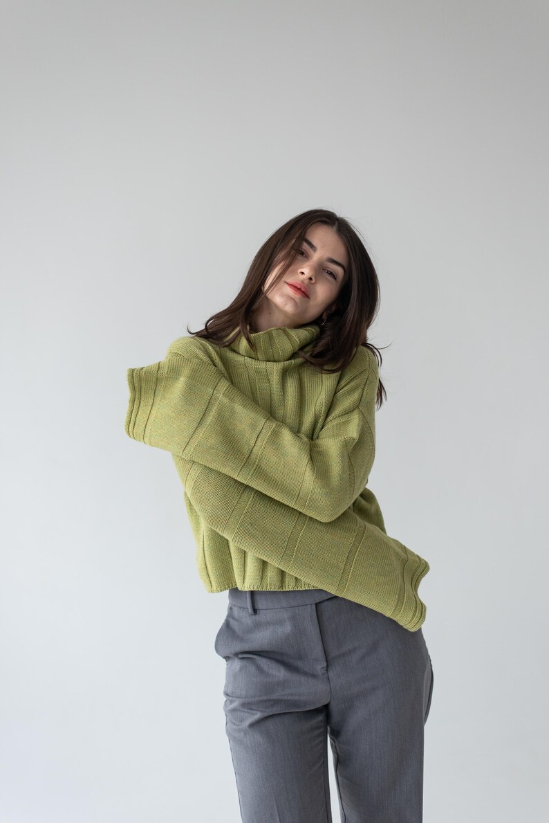 May include: A woman wearing a light green ribbed knit turtleneck sweater and gray trousers. The sweater has long sleeves and a high neckline. The pants are a solid gray color.