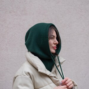 May include: A woman wearing a green knitted hooded neck warmer with a white puffer jacket. She is adjusting the strings of the neck warmer.
