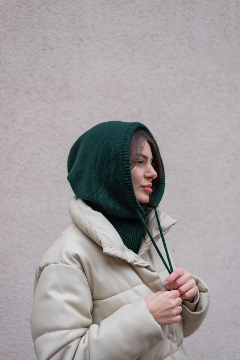 May include: A woman wearing a green knitted hooded neck warmer with a white puffer jacket. She is adjusting the strings of the neck warmer.