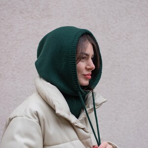 May include: A woman wearing a green knitted hooded neck warmer with a white puffer jacket. The neck warmer has a drawstring.