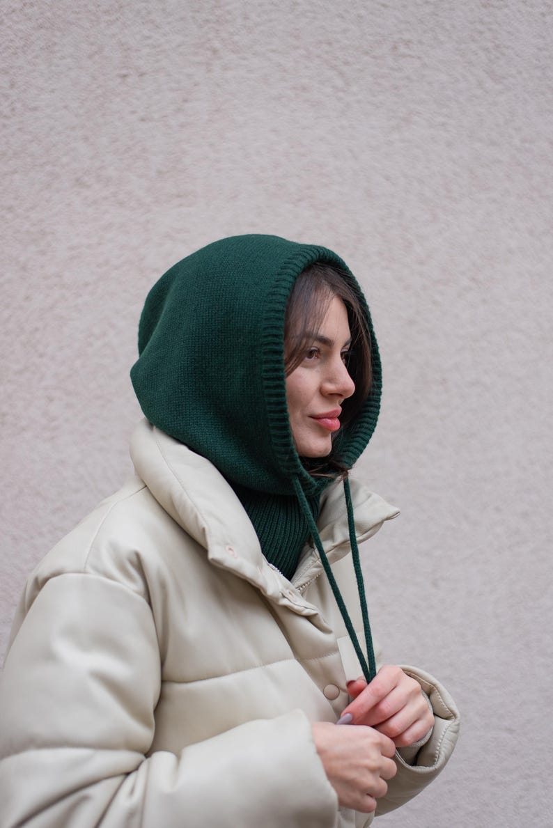 May include: A woman wearing a green knitted hooded neck warmer with a white puffer jacket. The neck warmer has a drawstring.